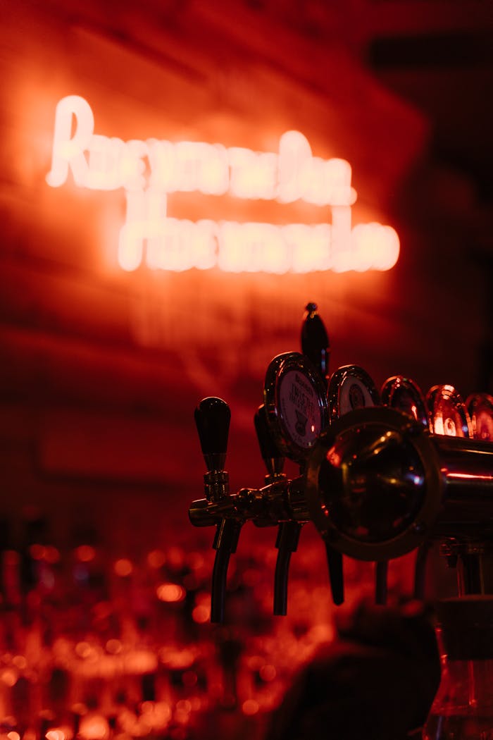 A close-up of beer taps with a blurred neon bar sign in the background.